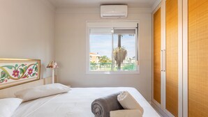 A bedroom with a bed, a window with a view of buildings and a palm tree, and a ceiling-mounted air conditioner. - Comfortable Beachside Apartment in Floripa Rvg101 (Florianópolis)