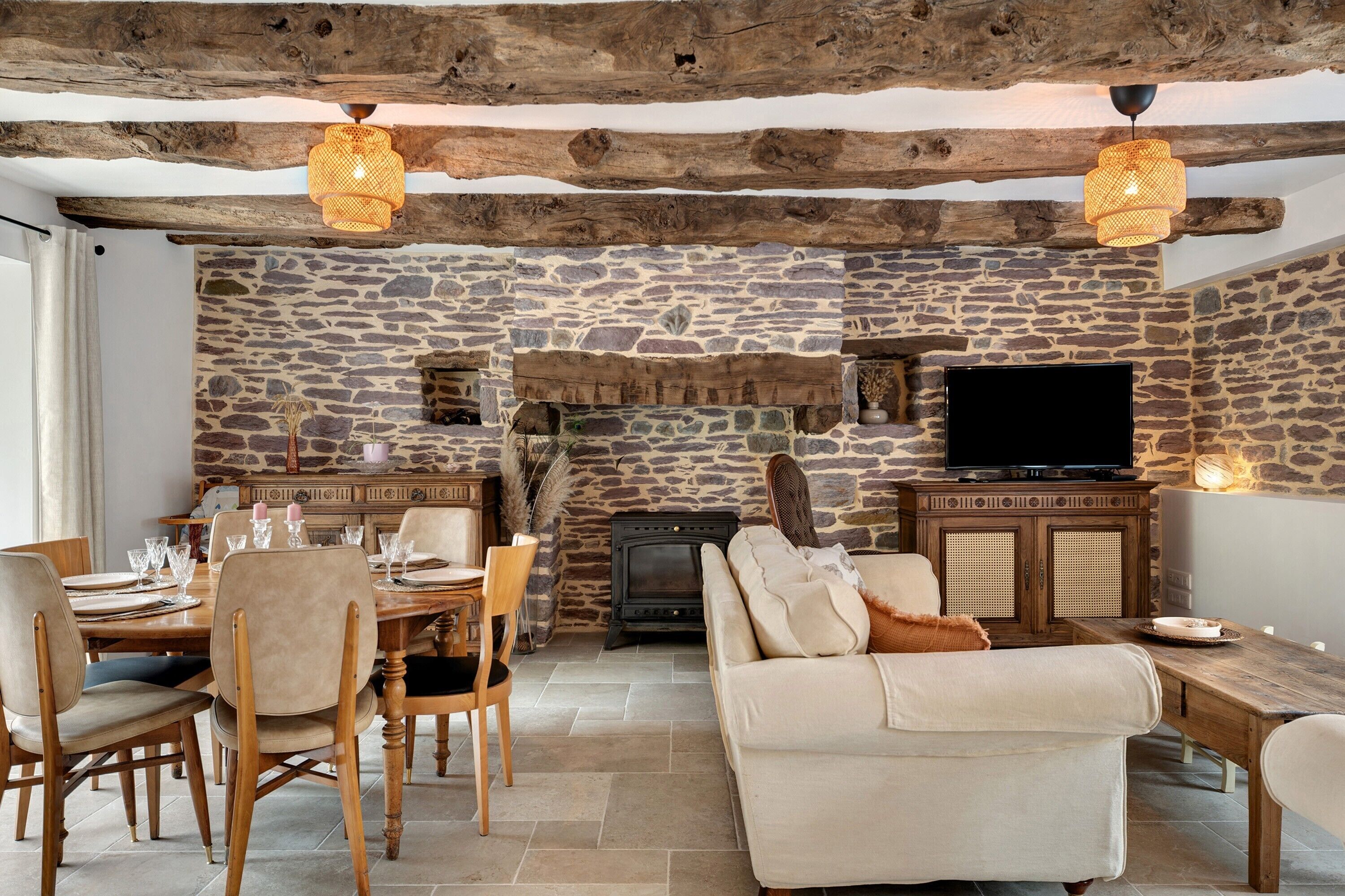 A rustic interior with a stone wall, wooden beams, a fireplace, a television, and a dining area.