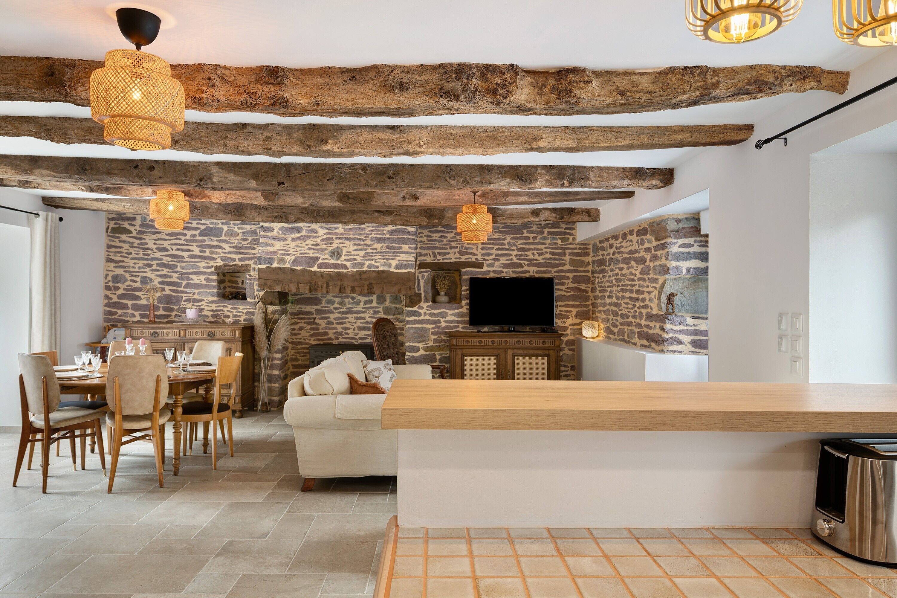Un intérieur rustique avec des poutres apparentes, des murs en pierre, un coin repas avec des chaises et une table, un canapé et un téléviseur fixé au mur.