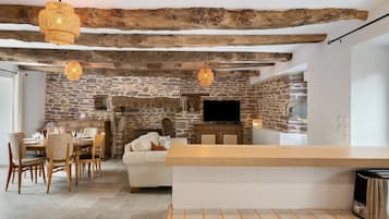 Un interior rústico con vigas de madera a la vista, paredes de piedra, una zona de comedor con sillas y una mesa, un sofá y un televisor montado en la pared.