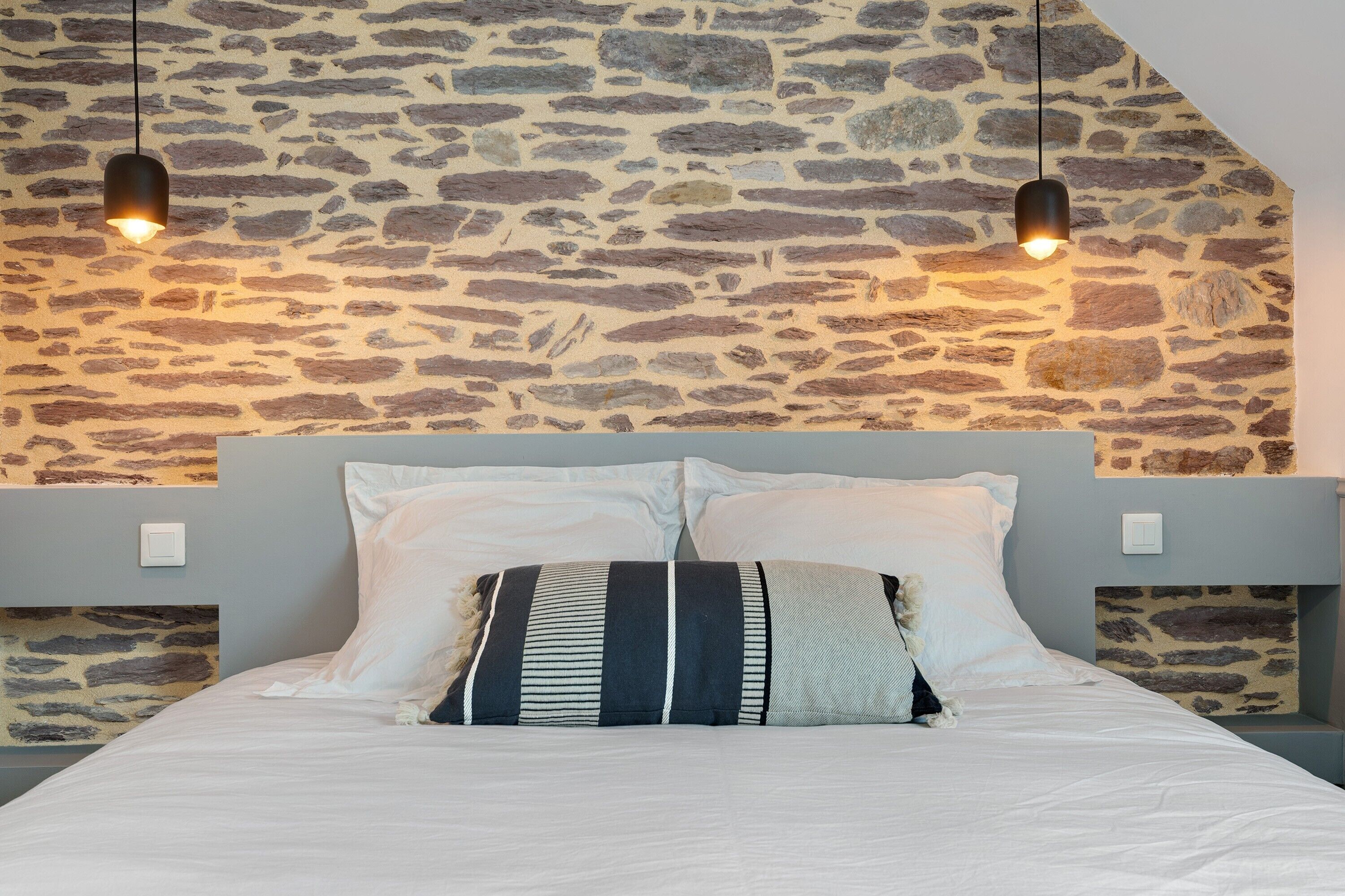 Cama con ropa de cama blanca y almohadas a rayas, frente a una pared de piedra con dos lámparas colgantes.