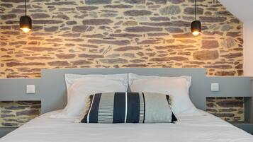 Cama con ropa de cama blanca y almohadas a rayas, frente a una pared de piedra con dos lámparas colgantes.