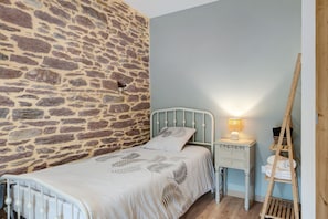 Un dormitorio con pared de piedra, una cama con edredón estampado con motivos de hojas, una mesita de noche con lámpara y una escalera de madera.