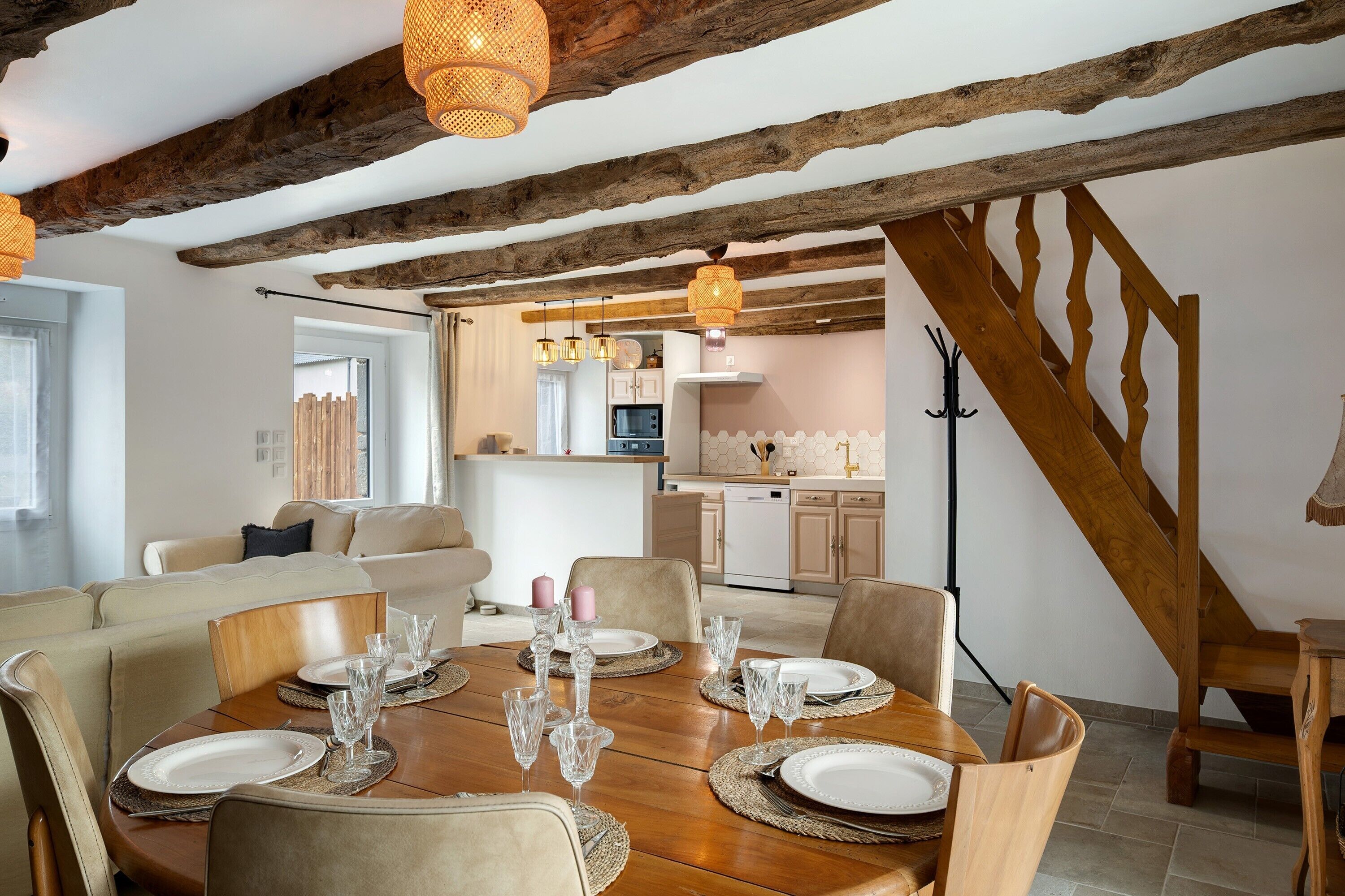 A dining area with a wooden table set for six, surrounded by chairs, in a room with exposed wooden beams and a kitchen in the background.