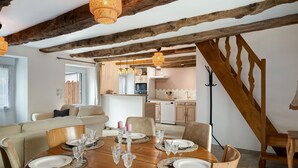 A dining area with a wooden table set for six, surrounded by chairs, in a room with exposed wooden beams and a kitchen in the background. - La Maison de Gautro – Holiday Home with Terrace and Garden (Tréhorenteuc)