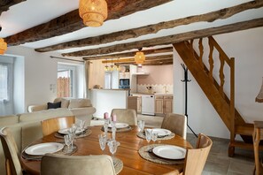 A dining area with a wooden table set for six, surrounded by chairs, in a room with exposed wooden beams and a kitchen in the background.