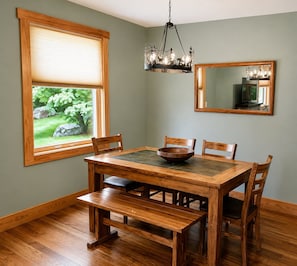A dining area with a wooden table and chairs, a chandelier, a window with a view of greenery, and a mirror on the wall.