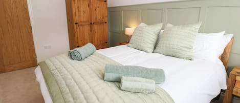 Una cama bien hecha con cubrecama acolchado, almohadas y toallas enrolladas. Se observa un armario de madera y una mesita de noche con una lámpara.