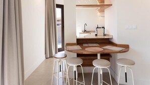 Eine kleine Küche mit Holzarbeitsplatte, Spüle und einer eingebauten Ablage für Gläser. Drei Hocker und ein Fenster mit Vorhängen sind ebenfalls vorhanden.