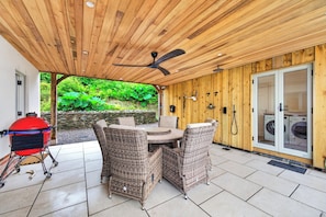 Eine Terrasse im Freien mit einem runden Tisch, Korbstühlen, einem Deckenventilator und einem Grill.