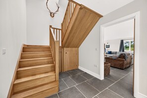 Un escalier en bois mène à un palier où se trouve une porte. De là, on a vue sur un salon avec un canapé et une fenêtre, ainsi qu’une décoration murale représentant une tête de cerf.