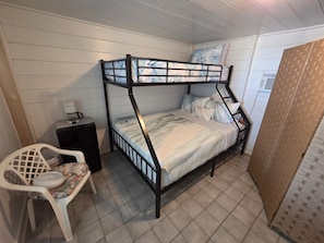 En våningssäng med en sänggavel, ett litet nattduksbord, en stol med ett blommigt sittkudd och en väggmonterad luftkonditioneringsenhet.