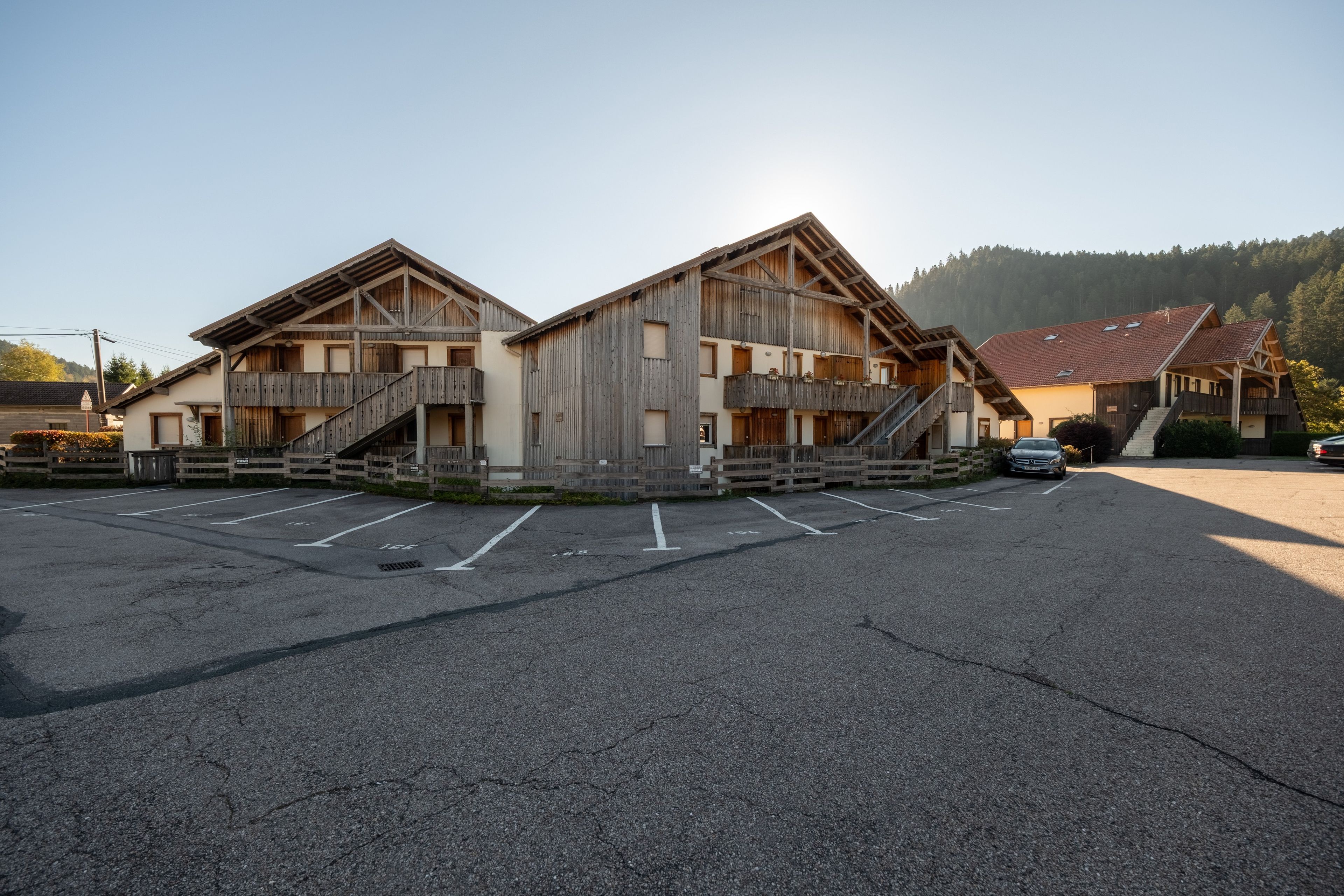 Ein zweigeschossiges Holzhaus mit einem Balkon im oberen Stockwerk, einem Parkplatz davor und einem bewaldeten Hügel im Hintergrund.