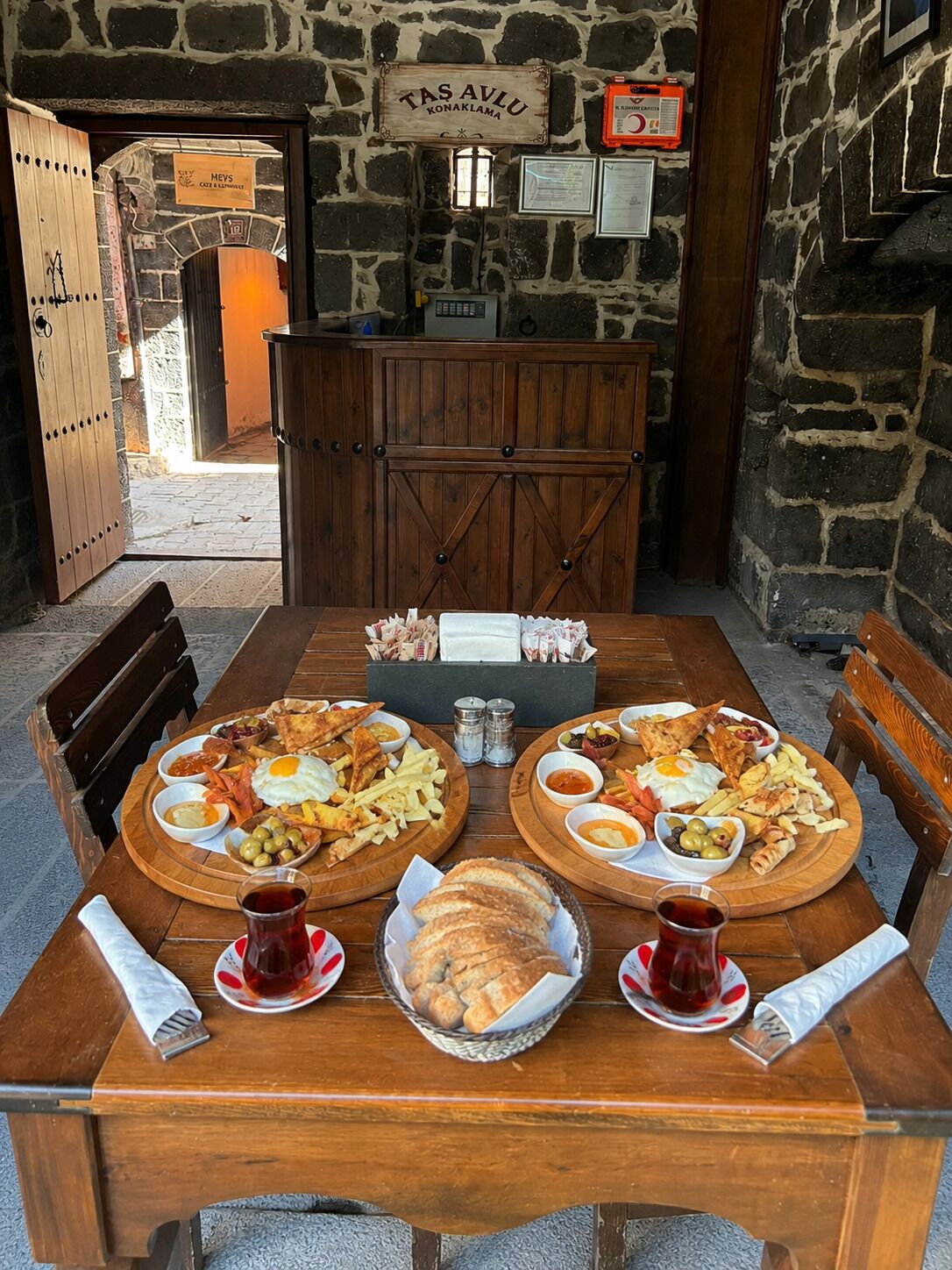 Una mesa de madera dispuesta con diversos alimentos para el desayuno, como huevos, salchichas y pan, en una habitación con paredes de piedra y un letrero que dice 'TAS AVLU KONAKLAMA'.
