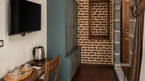 A narrow room with a brick wall, a television mounted on the wall, a wooden table with a kettle and cups, and a blue cabinet. - Taş Avlu Konaklama (Diyarbakir)