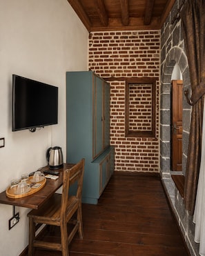 A narrow room with a brick wall, a television mounted on the wall, a wooden table with a kettle and cups, and a blue cabinet. - Taş Avlu Konaklama (Diyarbakir)