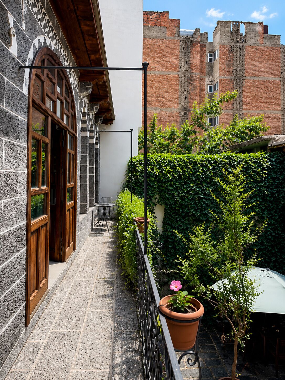 A narrow outdoor corridor with a stone wall on one side and a brick building on the other. There is a metal railing along the corridor, and a potted plant with a pink flower is visible.