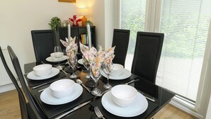 Mesa de jantar posta para quatro com pratos brancos, copos e talheres, rodeada por cadeiras pretas.