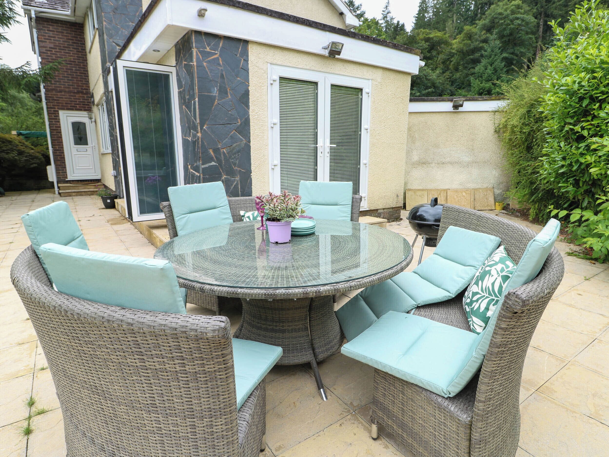 An outdoor patio with wicker furniture and a glass table, surrounded by greenery.