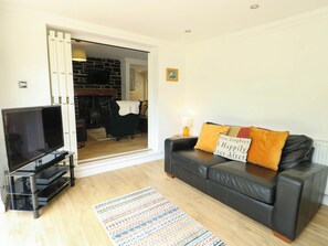 Ein Wohnzimmer mit einer schwarzen Ledersofa, einem Flachbildfernseher auf einem Standfuß, einem gemusterten Teppich und einer Tür, die in einen Küchenbereich führt.