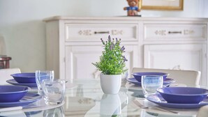 Una mesa de comedor con un jarrón blanco que contiene follaje verde y flores moradas, rodeada de platos azules y vasos transparentes.