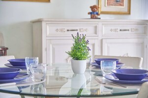 Una mesa de comedor con un jarrón blanco que contiene follaje verde y flores moradas, rodeada de platos azules y vasos transparentes.
