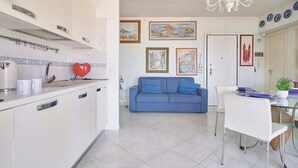 Una cocina moderna con armarios blancos, un objeto rojo con forma de corazón sobre la encimera, y un comedor con un sofá azul y sillas blancas.