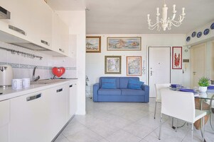Una cocina moderna con armarios blancos, un objeto rojo con forma de corazón sobre la encimera, y un comedor con un sofá azul y sillas blancas.