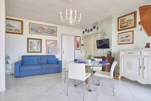 Una sala de estar con un sofá azul, una mesa de comedor con sillas, una araña y varias obras de arte enmarcadas en las paredes.