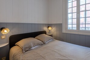 Quarto com cama, duas luminárias de cabeceira, janela com vista para edifícios e parede com lambris.