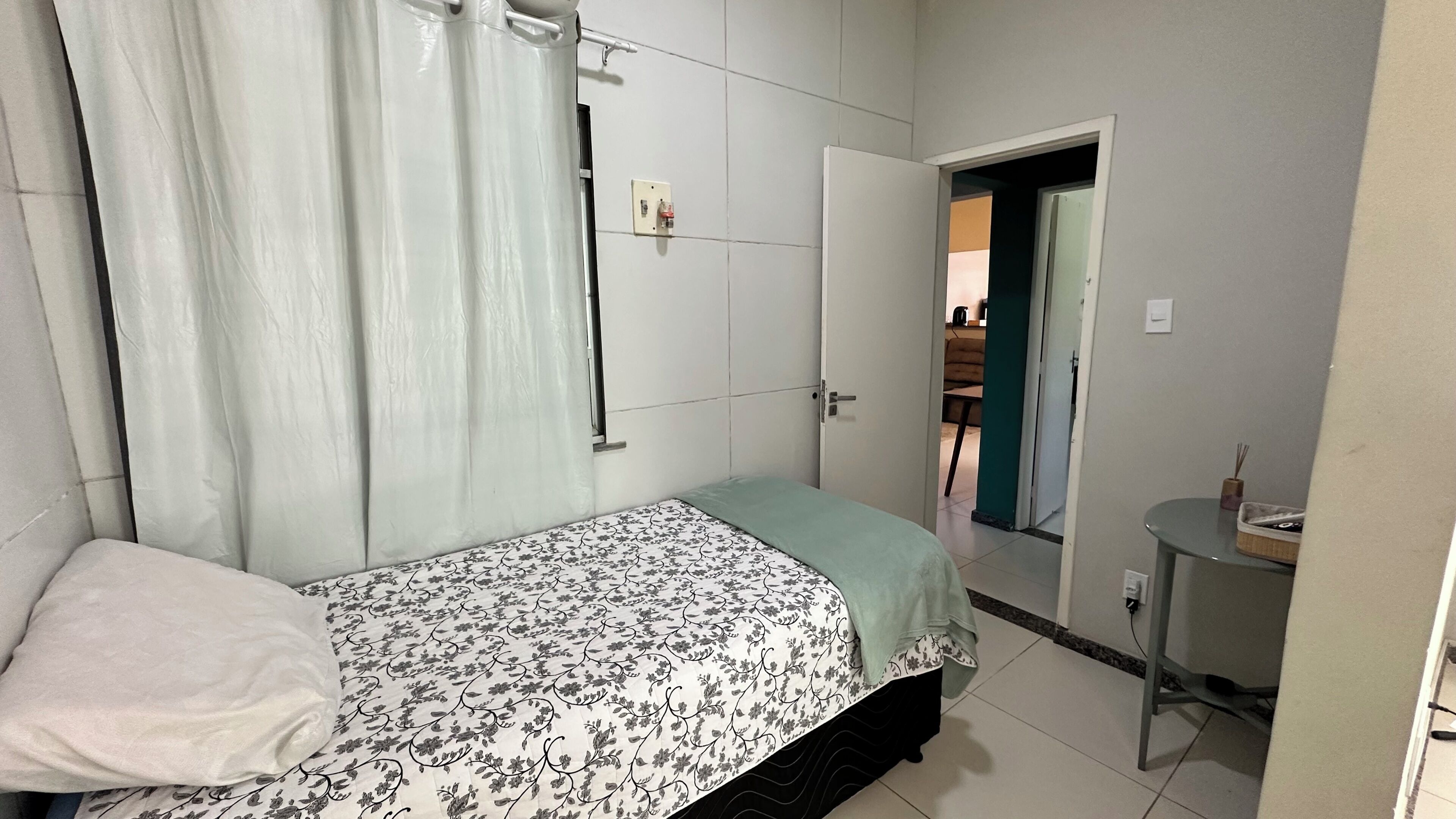 Un dormitorio con una cama, una mesita de noche, una cortina y una puerta que da a otra habitación.