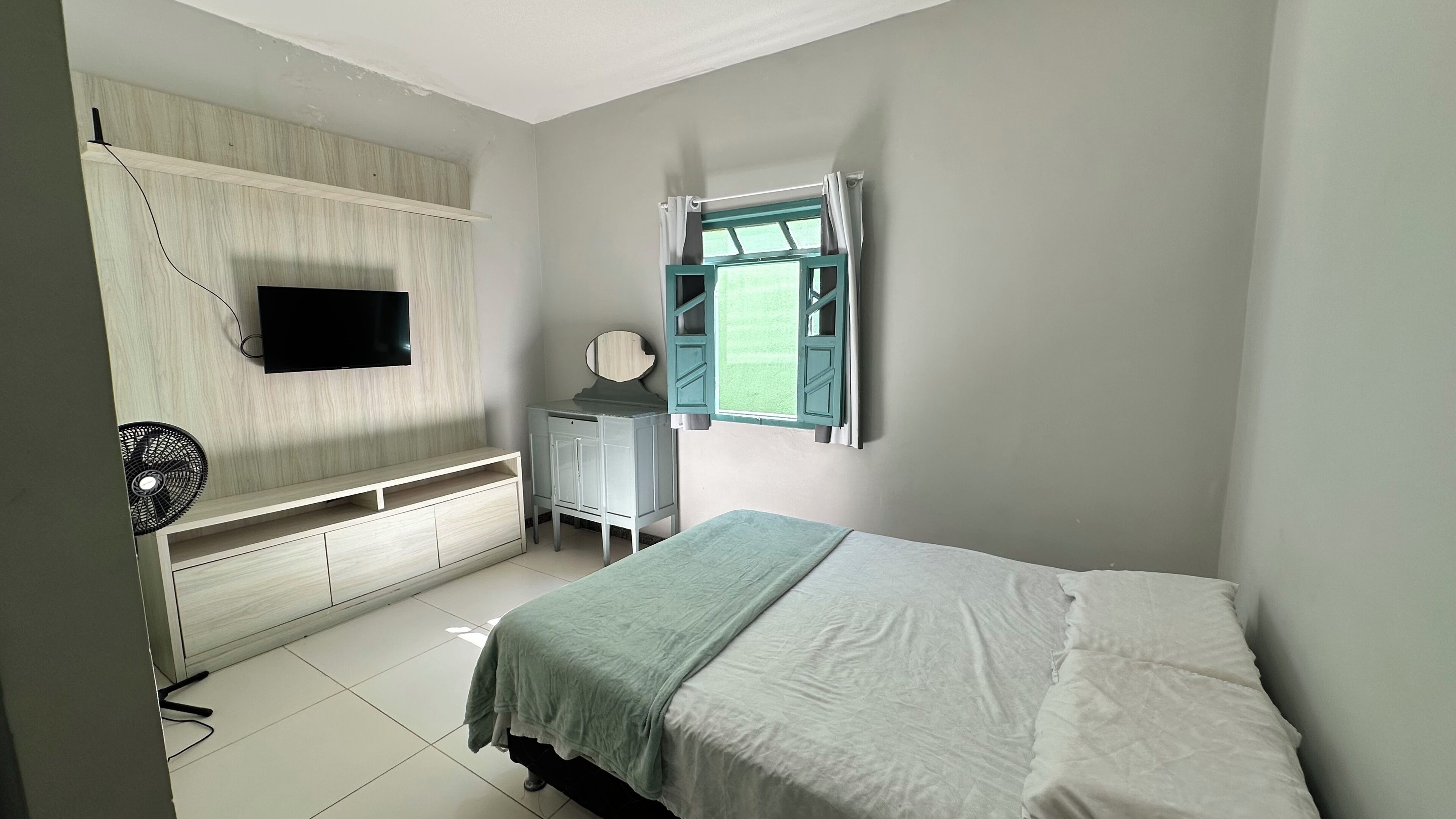 A bedroom with a bed, a TV mounted on the wall, a small desk with a chair, and a window with curtains.