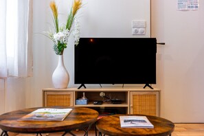 A living room with a flat-screen TV on a stand, two round wooden tables, a white vase with flowers, and a wall-mounted light switch. - Le Discret de Charles - Calme & Confort (Nancy)