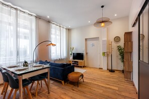 A modern living room with a dining area, wooden flooring, a blue sofa, a TV, and a clock on the wall. - Le Discret de Charles - Calme & Confort (Nancy)
