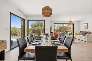 Ein Essbereich mit einem großen Tisch, gedeckt für sechs Personen, umgeben von Stühlen und mit Blick ins Freie durch große Fenster.