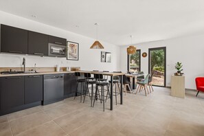 A modern kitchen with dark cabinets, a built-in oven, and a dining area with a wooden table and bar stools. - Maison de la Seranne - Avec piscine (Le Bosc)