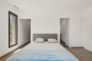 A modern bedroom with a large bed, two pillows, a grey headboard, and a view through a window. - Maison de la Seranne - Avec piscine (Le Bosc)