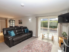 A living room with a black leather sofa, a colorful rug, a glass table, and a sliding glass door leading to a garden. - The Old Dairy (Liskeard)