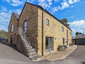 A stone building with a staircase leading to a door, a covered patio area, and a car parked in front. - The Old Dairy (Liskeard)