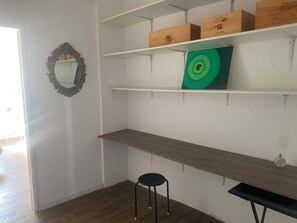 A room with white walls, a wooden floor, and a mirror on the left. There are three shelves with boxes and a green decorative item on the middle shelf. A black stool is in the corner. - Appartement climatisé avec vue mer et sur les toits du vieux Grasse (Grasse)