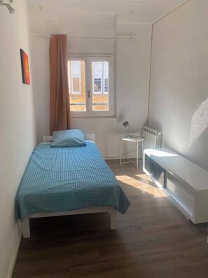 Ein kleines, helles Schlafzimmer mit einem Einzelbett, einem Heizkörper, einem kleinen Tisch und einem Fenster mit Vorhängen.