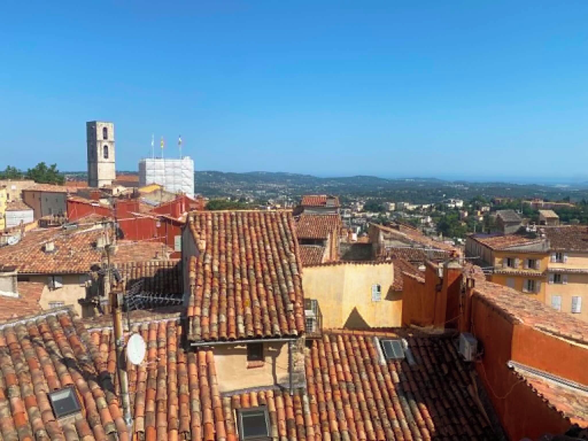 A cityscape with terracotta rooftops, a church tower, and a distant view of the coastline.