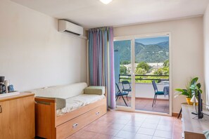 A room with a sofa, a small table, a chair, and a view of a balcony with a mountain backdrop. - Le Refuge de Rena - Appart Climatisé (San-Nicolao)