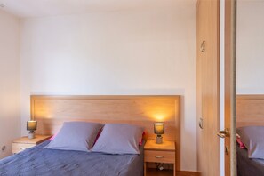 Et soveværelse med sengegavl i træ, to sengeborde med lamper, en seng med gråt dyne og en garderobe i træ med spejl.