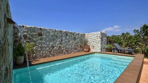 Ein rechteckiger Pool mit einer Steinwand im Hintergrund, einer Holzterrasse und Liegestühlen.