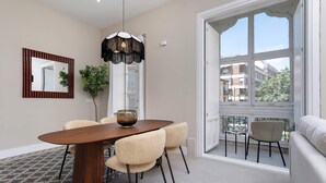 Un espace repas comprenant une table en bois, des chaises, un luminaire suspendu et une grande fenêtre donnant sur des immeubles.