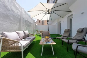 Un espacio al aire libre iluminado por el sol con sofás, sillones, una mesa con bebidas y una sombrilla grande.