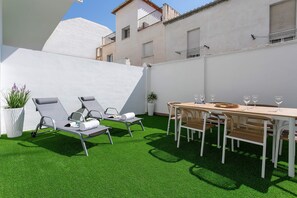 Un patio con césped artificial, una mesa lista para comer y sillones de descanso.