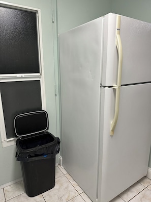 Eine Küche mit einem Edelstahl-Kühlschrank, einer schwarzen Mülltonne und einem Fenster mit schwarzem Fliegengitter.