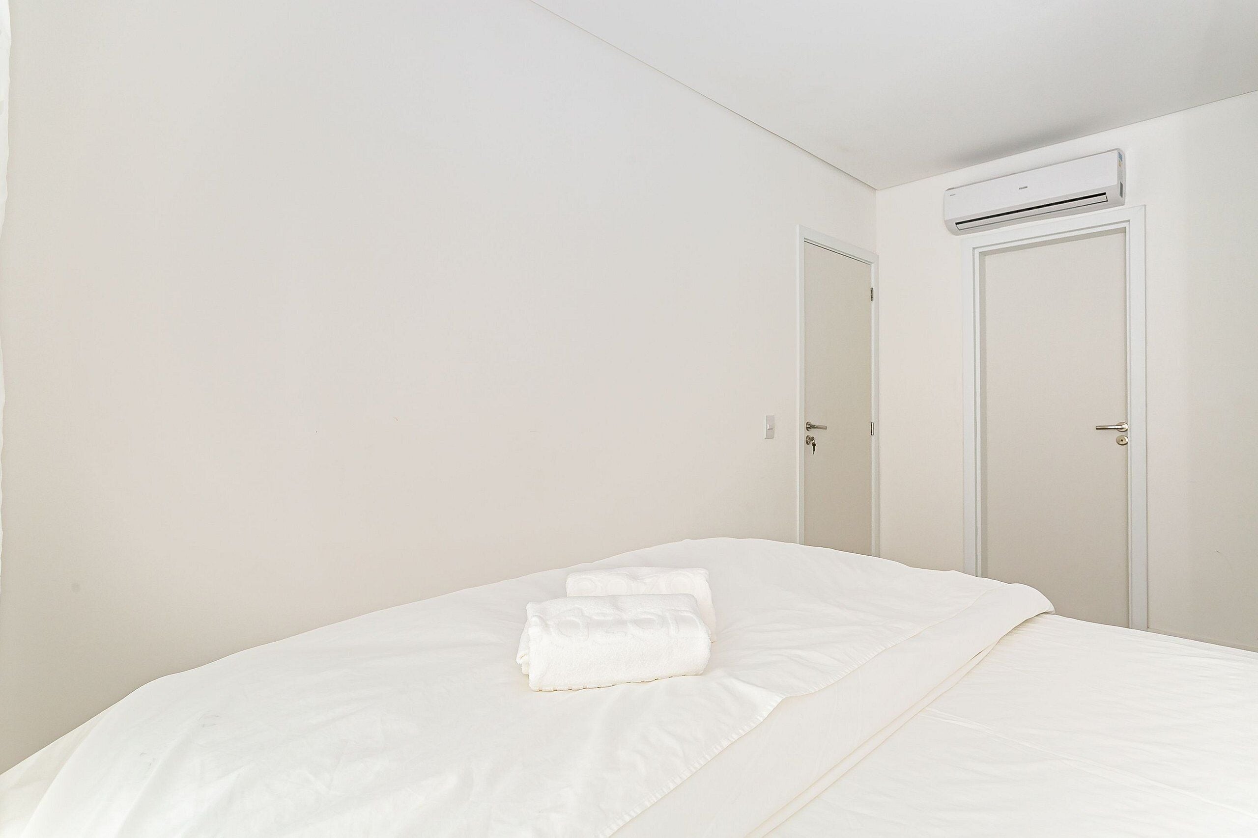 A clean, white room with a bed, a door, and an air conditioning unit.